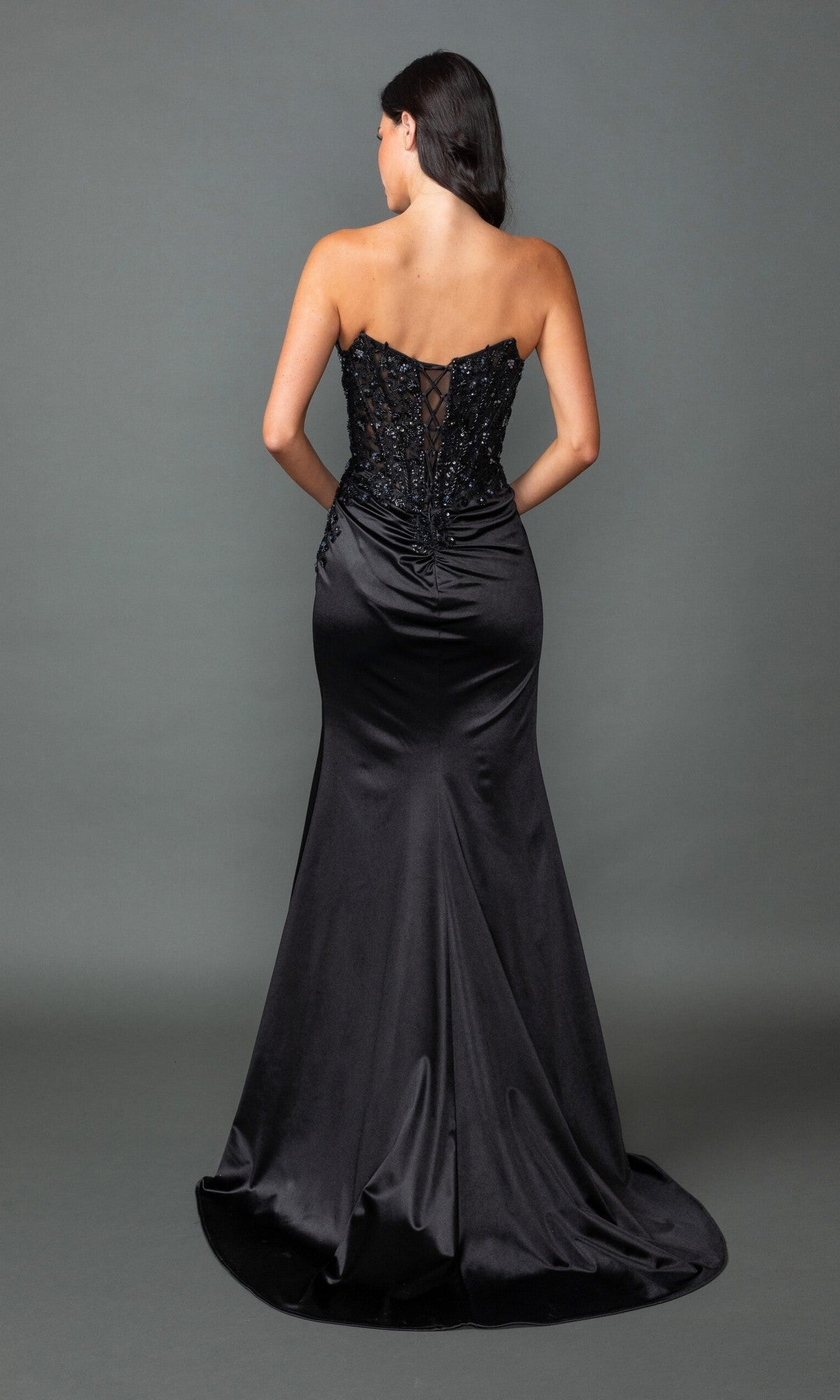 A woman faces away from the camera in the Lace-Up Long Black Prom Dress: Dancing Queen 4555, featuring a strapless design, sweetheart corset bodice, beaded embroidery, lace-up back, and a flowing satin skirt.